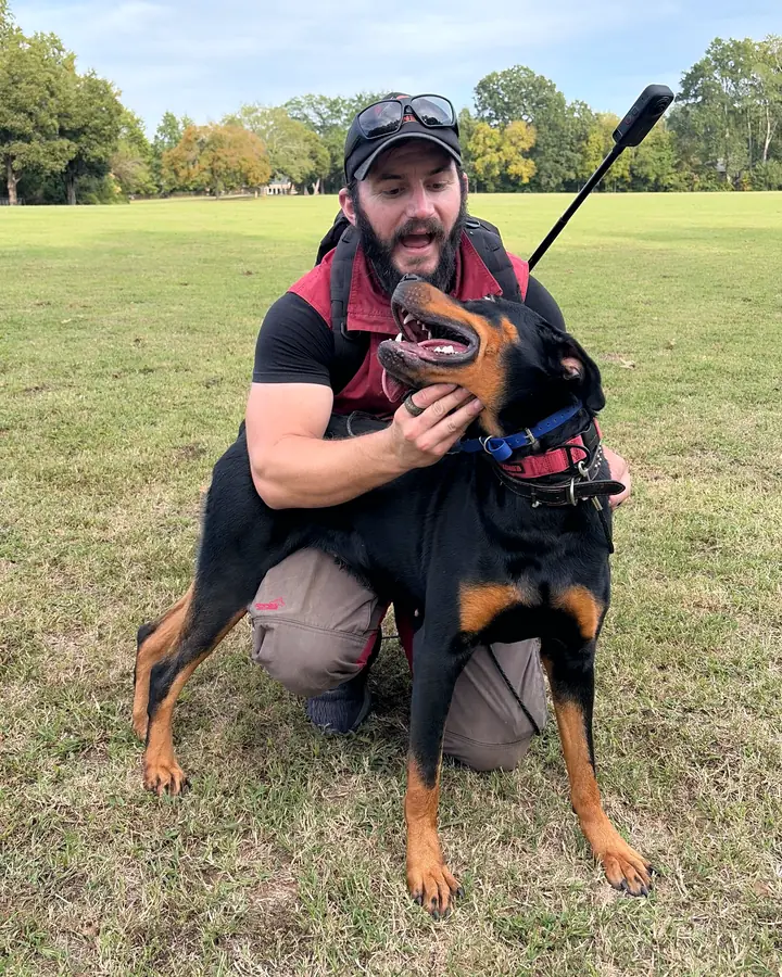Kelton, Angier K9’s head trainer, kneeling in a park and laughing as he hugs a black-and-tan dog who looks up at him