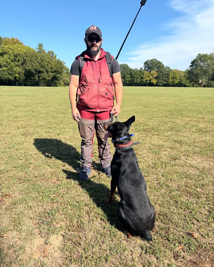 Kelton standing in an open field with a dog sitting close, the backpack-mounted 360° camera on its pole above him