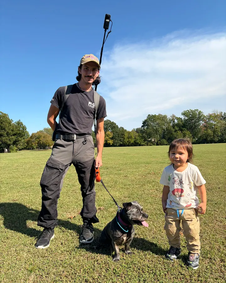 Christian Scharphorn, Angier K9’s founder, standing in a park with a small dark dog on a leash and his young daughter at his side